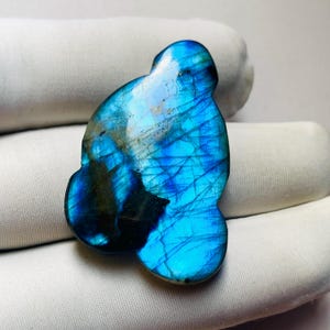 Peut inclure: Une pierre précieuse de labradorite polie, présentant une irisation bleue et turquoise saisissante. La pierre a une forme organique et présente un jeu de lumière, soulignant sa beauté naturelle. La pierre est posée sur un fond blanc.
