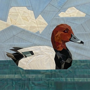 May include: A fabric quilt block featuring a geometric, pieced design of a duck with a reddish-brown head, black chest, and white body. The background consists of light blue sky segments with white clouds and dark teal water ripples below.