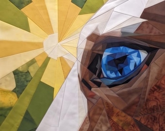 Może przedstawiać: Tekstylna dekoracja ścienna typu patchwork przedstawiająca zbliżenie na kocie oko w odcieniach brązu i czerni. Oko ma żywy, głęboki niebieski kolor. Za kotem znajduje się wzór promieni słonecznych wykonany z żółtych i zielonych segmentów tkaniny.