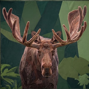 May include: A fabric wall hanging featuring a detailed moose portrait constructed from various brown and tan textured fabric pieces. The moose has large, prominent antlers and is set against a geometric background of layered green fabric.