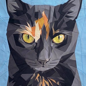 Könnte beinhalten: Ein geometrischer Quilt mit einem Katzengesicht. Die Katze hat gelbe Augen, eine schwarze Nase und eine Mischung aus schwarzen, grauen und orangefarbenen Flecken. Der Hintergrund ist hellblau und der Quilt besteht aus Stoffstücken.