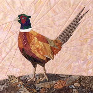 May include: A fabric quilt wall hanging featuring a pieced pheasant with a green head, red eye patch, white neck ring, and brown patterned body. The bird stands on a textured ground of brown fabric scraps against a pale pink sunburst background.