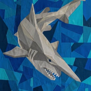 May include: A fabric quilt featuring a geometric shark design composed of various shades of grey, white, and blue. The shark is depicted in a swimming motion against a background of angular, multi-tonal blue fabric pieces arranged in a mosaic pattern.
