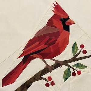 May include: A patchwork design of a red cardinal bird perched on a brown branch with green leaves and red berries. The bird has a black mask and a crest. The background is a light beige colour.
