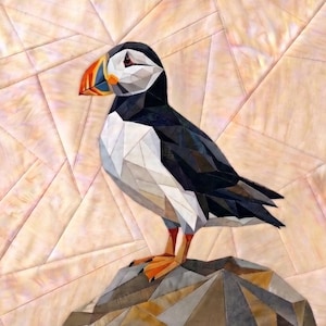 Könnte beinhalten: Ein geometrisches Stoffkunstwerk, das einen Papageientaucher mit schwarz-weiß-orangem Schnabel, schwarz-weißem Gefieder und orangen Füßen zeigt. Der Vogel steht auf einer grauen, geometrischen Felsformation vor einem Hintergrund aus hellpfirsichfarbenen Stoffdreiecken.