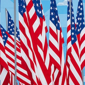 Op de afbeelding: Een levendige afbeelding met meerdere Amerikaanse vlaggen die wapperen tegen een lichtblauwe achtergrond. De vlaggen hebben rode en witte strepen met een blauw kanton vol witte sterren. De vlaggen zijn in een dynamische compositie gerangschikt.