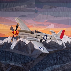 Könnte beinhalten: Ein Stoffquilt, der ein silbernes P-51 Mustang Kampfflugzeug im Flug über schneebedeckten Bergen darstellt. Das Flugzeug hat eine rot-gelb karierte Nase, den Schriftzug B6 P auf dem Rumpf sowie blau-weiße Sternabzeichen auf den Tragflächen.