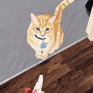 May include: A foundation paper pieced quilt block featuring an orange tabby cat with a blue collar tag, positioned on a gray background. Below the cat, a small white mouse toy with a red polka-dot heart tail rests on a brown wood-grain fabric.