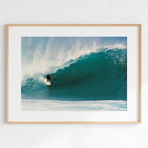 Könnte beinhalten: Ein gerahmter Kunstdruck, der einen Surfer in einer großen, sich kräuselnden türkisfarbenen Meereswelle zeigt. Der Surfer befindet sich auf einem weißen Surfbrett, umgeben von tiefblauem Wasser und weißer Gischt, in einem hellen Holzrahmen.