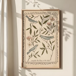 Op de afbeelding: Een ingelijste kunstdruk met drie blauwe vogels die zich bevinden tussen groene bladeren en roze bloemen. Het kunstwerk bevat de tekst "consider the lilies in the field" in een decoratieve lijst.