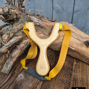 May include: A wooden slingshot with yellow rubber bands and a black leather pouch. The slingshot has a natural wood finish and the initials "RK" are carved into the handle.