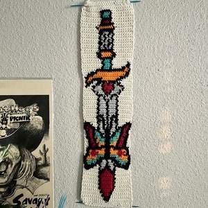 Könnte beinhalten: Ein weißer gehäkelter Wandbehang mit einem bunten Pixel-Design eines Dolches mit einem Schmetterling auf der Klinge. Der Dolch ist rot, schwarz, blau und orange. Der Schmetterling ist gelb, orange, blau und rot.