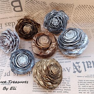 May include: An assortment of decorative pine cone flowers in various metallic colors, including silver, gold, and bronze. The flowers are arranged on a newspaper background. The text "Nature Treasures By Eli" is visible.