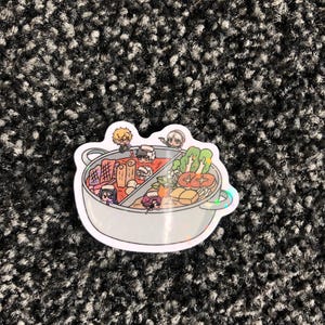 May include: A sticker featuring a divided pot of food with cartoon characters. The pot contains various ingredients, including vegetables and meat. The sticker has a holographic border and is placed on a textured, dark gray surface.