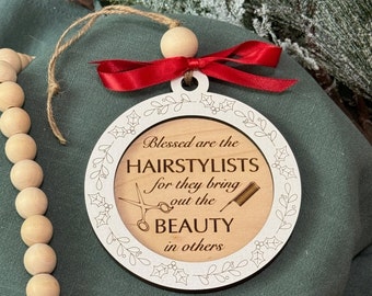 May include: A wooden Christmas ornament with a white border and a message that reads "Blessed are the hairstylists for they bring out the beauty in others". The ornament is decorated with a red ribbon and a wooden bead.