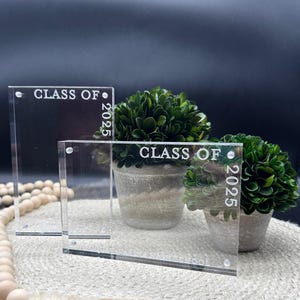 Könnte beinhalten: Zwei transparente Acryl-Bilderrahmen mit dem eingravierten Text "Class of 2025". Die Rahmen stehen auf einer gewebten Oberfläche mit kleinen Topfpflanzen davor.
