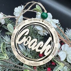 May include: Wooden ornament with the word "Coach" in a decorative font. The ornament is surrounded by a circle with the words "Competitive, Dedicated, Leader, Role Model, Passionate, Mentor, Admirable, Inspirational, Supportive, Hard Working" inscribed around it.