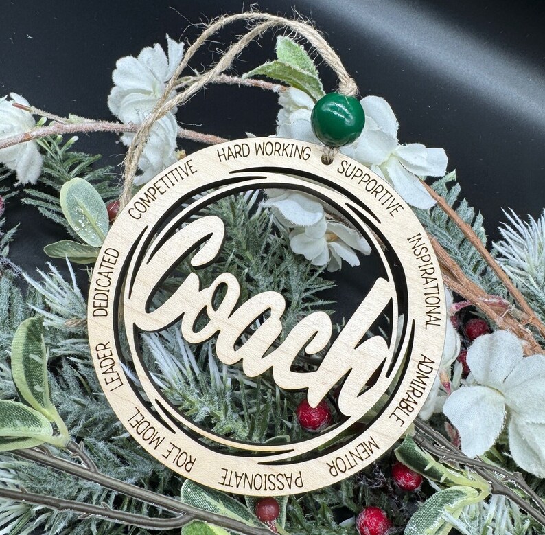 May include: Wooden ornament with the word "Coach" in a decorative font. The ornament is surrounded by a circle with the words "Competitive, Dedicated, Leader, Role Model, Passionate, Mentor, Admirable, Inspirational, Supportive, Hard Working" inscribed around it.