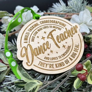 Puede incluir: Un adorno de madera con fondo marr&oacute;n y texto dorado. El adorno dice "A multi-tasking, choreographing rock star who lives to inspire and loves to encourage. They're kind of a big deal." El adorno tiene forma de c&iacute;rculo y tiene una cinta verde atada.