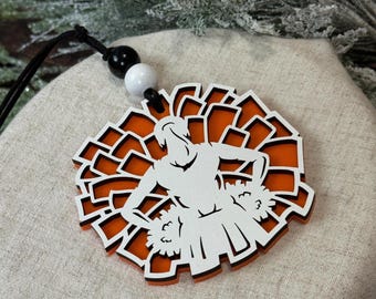 May include: Orange and white wooden ornament featuring a silhouette of a cheerleader holding pom-poms. The ornament is attached to a black and white cord.