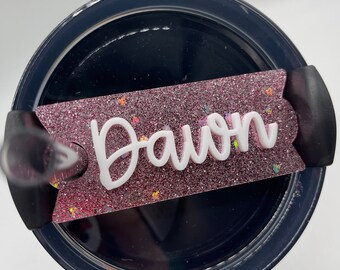 Puede incluir: Una tapa de vaso rosa con purpurina con una etiqueta de nombre blanca que dice "Daivon".