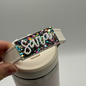 Peut inclure: Une tasse blanche personnalisée avec une étiquette de nom en paillettes colorées qui dit "Sara".