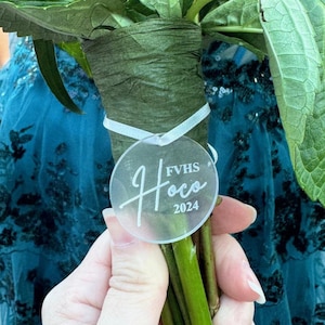 Puede incluir: Un ramo de flores con tallos verdes envuelto en papel verde, atado con una cinta blanca. Una etiqueta redonda transparente con la palabra "Hoco" y "FVHS 2024" en escritura blanca está adjunta al ramo.