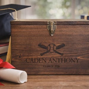 May include: A wooden keepsake box with a baseball theme, featuring a baseball and crossed bats design. The box is engraved with the name "CADEN ANTHONY" and "CLASS OF 2026". A graduation cap, books, and a diploma with a red ribbon are also in the image.