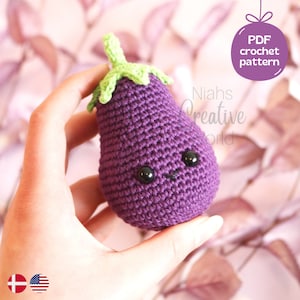 Puede incluir: Un juguete amigurumi de berenjena tejido a crochet con un tallo verde y una cara linda. El juguete se está sosteniendo en una mano contra un fondo rosa y verde. El texto "PDF crochet pattern" es visible en la esquina superior derecha de la imagen.