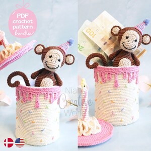 May include: A crocheted birthday cake with a monkey wearing a party hat peeking out of the top. The cake is decorated with sprinkles and pink frosting. The cake is a money box with a removable lid.