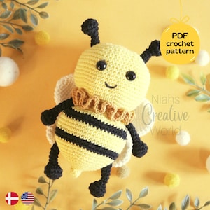 May include: A crocheted bee toy with black and yellow stripes, black eyes, and a brown crocheted collar. The bee is sitting on a yellow surface with green leaves and white and yellow balls.