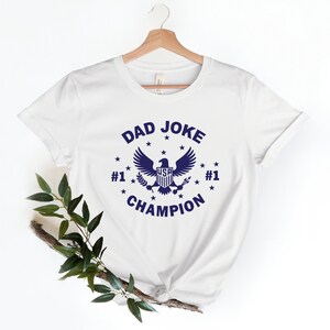 Op de afbeelding: Wit T-shirt met een blauwe afbeelding van een adelaar met de tekst "DAD JOKE #1 CHAMPION #1" eromheen.