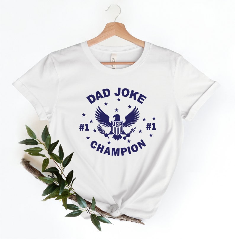 Op de afbeelding: Wit T-shirt met een blauwe afbeelding van een adelaar met de tekst "DAD JOKE #1 CHAMPION #1" eromheen.