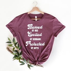 K&ouml;nnte beinhalten: Burgunderfarbenes T-Shirt mit wei&szlig;em Text, der "Gesegnet von Gott, Verw&ouml;hnt vom Ehemann, Gesch&uuml;tzt von beiden" lautet.