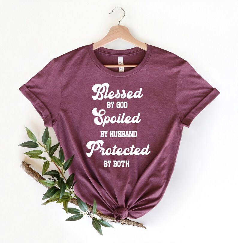 K&ouml;nnte beinhalten: Burgunderfarbenes T-Shirt mit wei&szlig;em Text, der "Gesegnet von Gott, Verw&ouml;hnt vom Ehemann, Gesch&uuml;tzt von beiden" lautet.