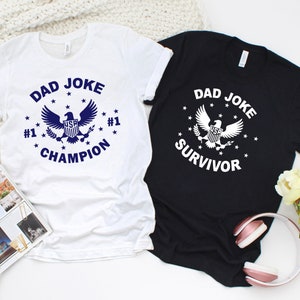 Op de afbeelding: Twee witte T-shirts met een blauwe adelaar afbeelding en de tekst "Dad Joke Champion" en "Dad Joke Survivor".