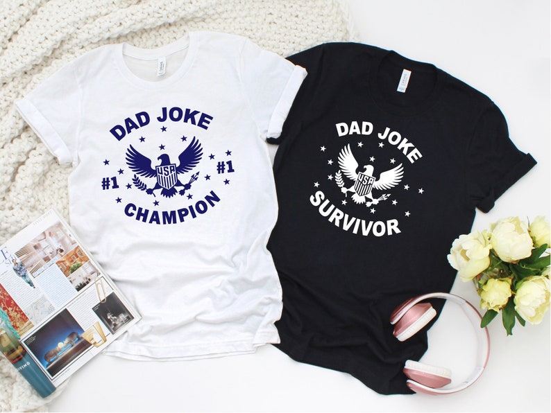 Op de afbeelding: Twee witte T-shirts met een blauwe adelaar afbeelding en de tekst "Dad Joke Champion" en "Dad Joke Survivor".
