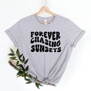 May include: A gray t-shirt with the text "FOREVER CHASING SUNSETS" printed in black. The text is in a retro, bubbly font.