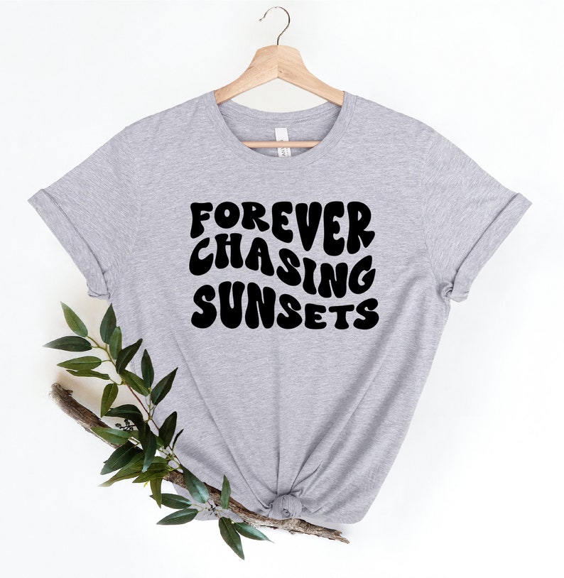 May include: A gray t-shirt with the text "FOREVER CHASING SUNSETS" printed in black. The text is in a retro, bubbly font.