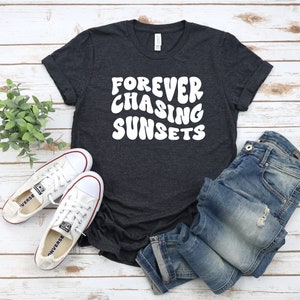 May include: A dark grey t-shirt with the text "FOREVER CHASING SUNSETS" in white, styled with blue jeans and white sneakers.