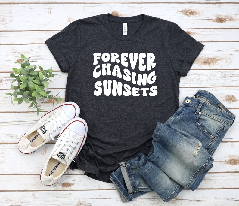 May include: A dark grey t-shirt with the text "FOREVER CHASING SUNSETS" in white, styled with blue jeans and white sneakers.