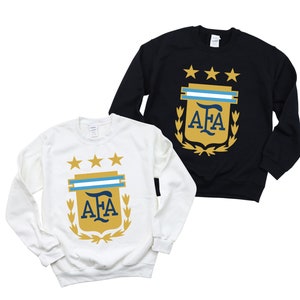 May include: Two long-sleeved sweatshirts with the Argentinian Football Association (AFA) logo. The sweatshirt on the left is white with a gold logo. The sweatshirt on the right is black with a gold logo.