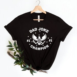 Op de afbeelding: Zwart T-shirt met een witte afbeelding van een adelaar met de tekst "Dad Joke Champion" en "#1" aan weerszijden van de adelaar.