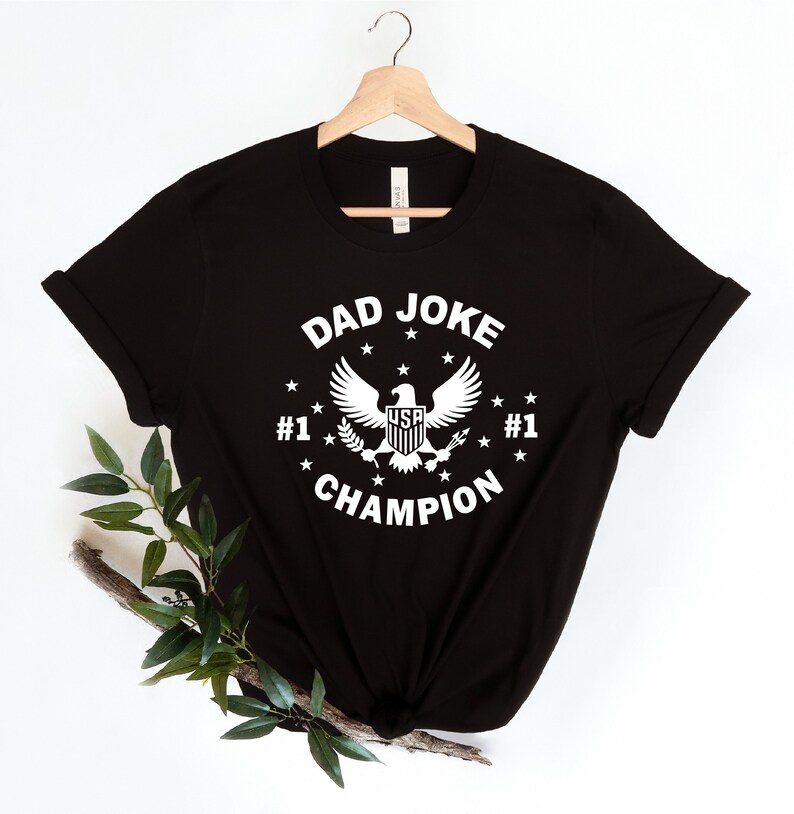 Op de afbeelding: Zwart T-shirt met een witte afbeelding van een adelaar met de tekst "Dad Joke Champion" en "#1" aan weerszijden van de adelaar.