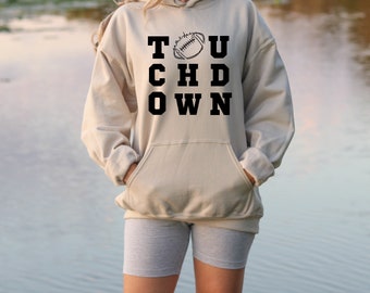 Op de afbeelding: Een beige hoodie met een zwarte grafische afbeelding waarop "TOUCHDOWN" staat met een voetbal in het midden van de "O".