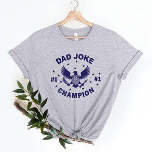 Op de afbeelding: Grijs T-shirt met een blauwe afbeelding van een adelaar met de tekst "DAD JOKE #1 CHAMPION #1" en sterren rond de adelaar.