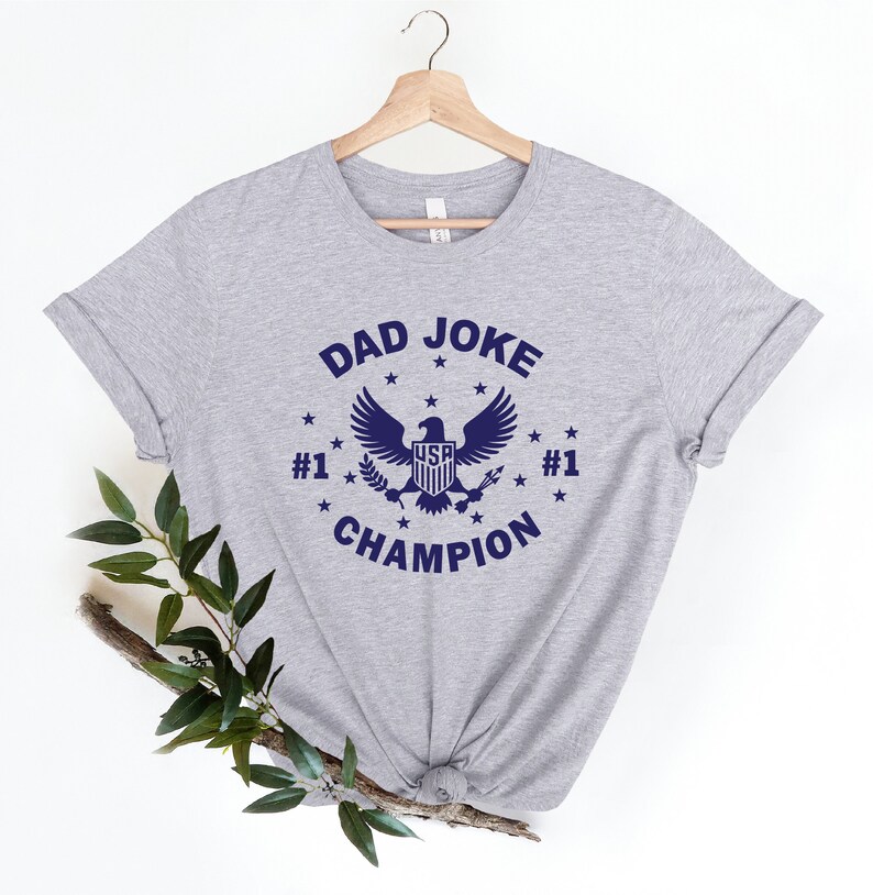 Op de afbeelding: Grijs T-shirt met een blauwe afbeelding van een adelaar met de tekst "DAD JOKE #1 CHAMPION #1" en sterren rond de adelaar.