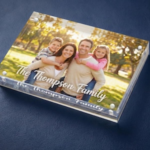 Peut inclure: Un bloc photo en acrylique transparent avec un portrait de famille et le texte "The Thompson Family" en écriture blanche. L'image montre une famille de quatre personnes dans un cadre de parc.