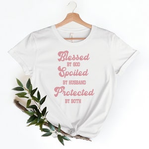 K&ouml;nnte beinhalten: Wei&szlig;es T-Shirt mit einer pinkfarbenen Grafik, auf der in einer Retro-Schrift "Gesegnet von Gott, Verw&ouml;hnt vom Ehemann, Gesch&uuml;tzt von beiden" steht.