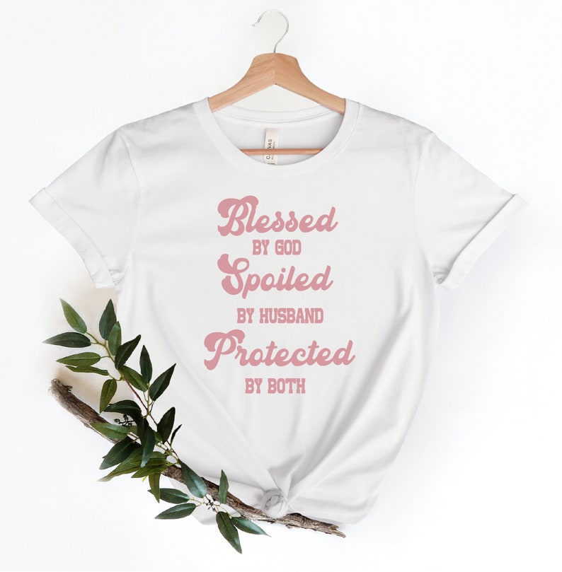 K&ouml;nnte beinhalten: Wei&szlig;es T-Shirt mit einer pinkfarbenen Grafik, auf der in einer Retro-Schrift "Gesegnet von Gott, Verw&ouml;hnt vom Ehemann, Gesch&uuml;tzt von beiden" steht.